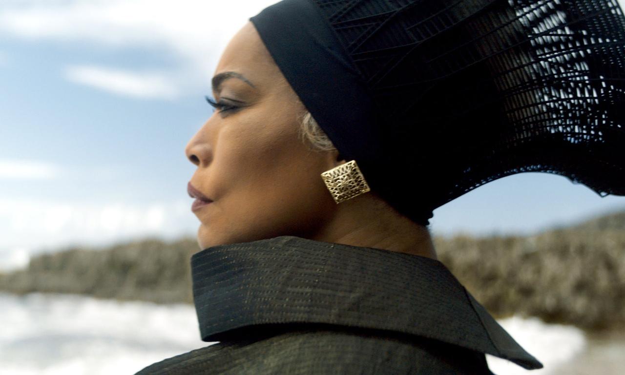 Still image of a woman wearing a headdress and square gold earrings. She is looking towards the ocean