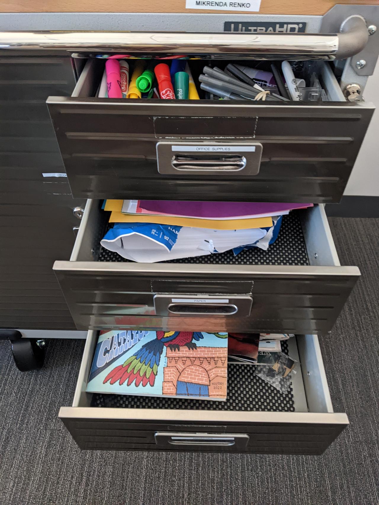 A photograph of a gunmetal grey cart with three open drawers featuring magazines, collaging materials, markers